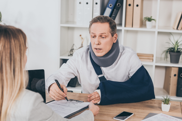 Worker In Neck Brace And Arm Bandage At Table Pointing With Pen